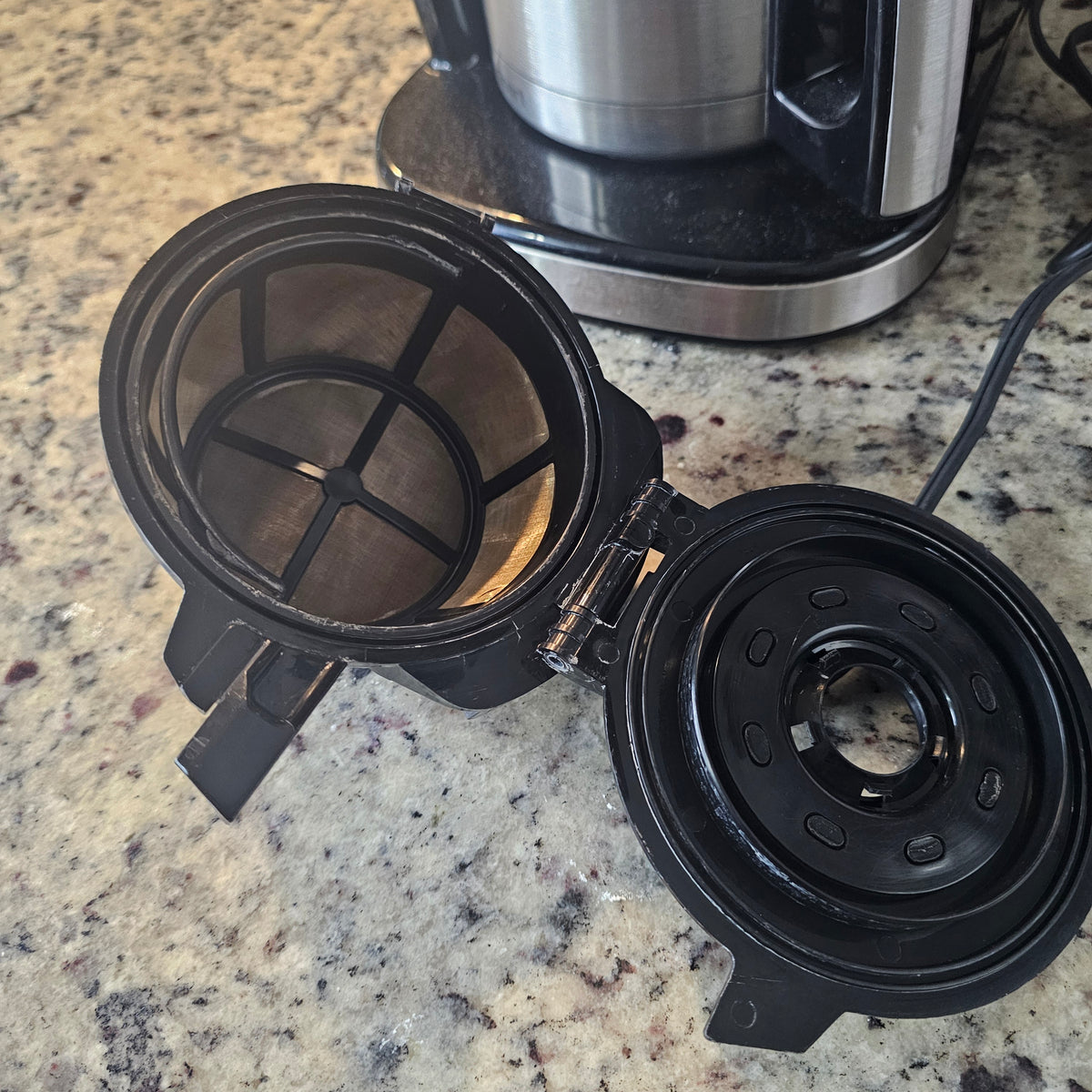 Open black coffee maker with a round mesh filter on a speckled countertop.