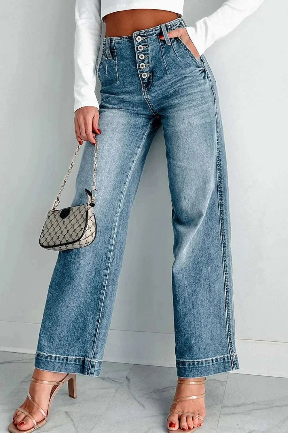 Woman wearing button fly high waist jeans with wide legs and light wash denim, paired with white top and beige heels, holding a small patterned handbag.