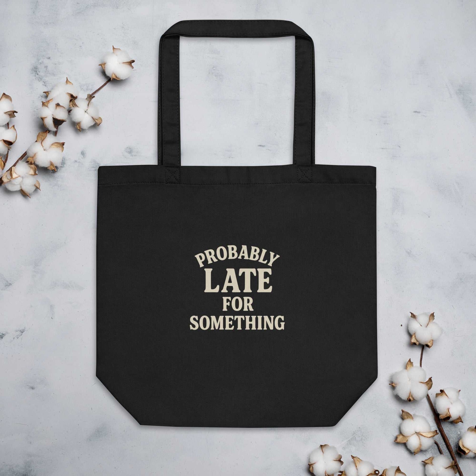 Black organic cotton eco tote bag with "Probably Late for Something" text and dual straps surrounded by cotton branches on a light background
