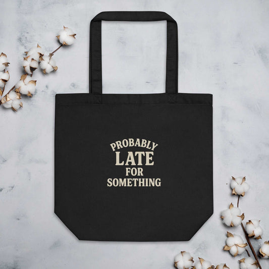 Black organic cotton eco tote bag with "Probably Late for Something" text and dual straps surrounded by cotton branches on a light background